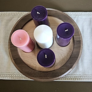 May include: A wooden candle holder with a circular design, featuring five pillar candles. The candles are in shades of pink, white, and purple. The holder is placed on a neutral-colored table runner.