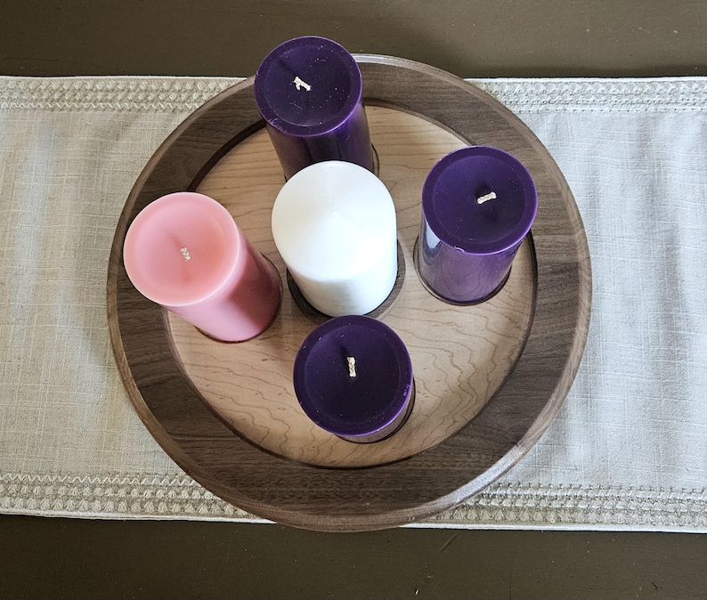 May include: A wooden candle holder with a circular design, featuring five pillar candles. The candles are in shades of pink, white, and purple. The holder is placed on a neutral-colored table runner.
