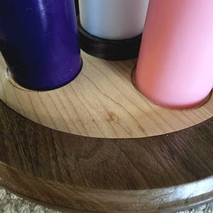 May include: A wooden candle holder with three candles. The candles are purple, white, and pink. The holder is made of light-colored wood with a dark brown trim. The candles are cylindrical and stand upright in the holder.