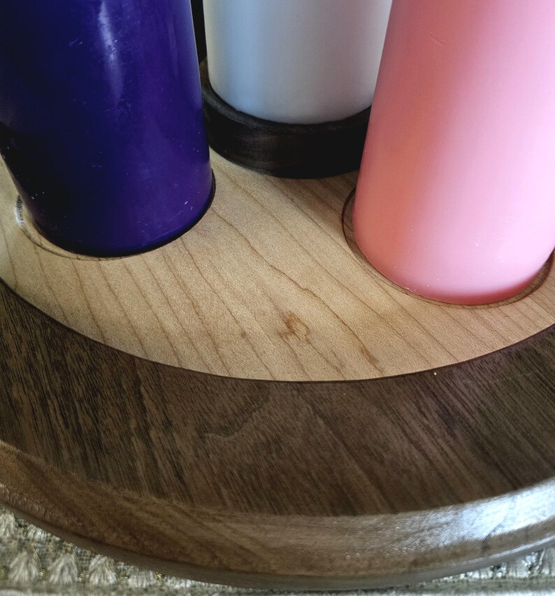 May include: A wooden candle holder with three candles. The candles are purple, white, and pink. The holder is made of light-colored wood with a dark brown trim. The candles are cylindrical and stand upright in the holder.
