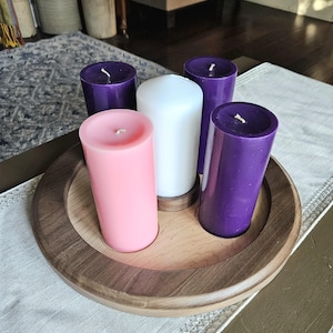 May include: A wooden candle holder with five pillar candles. The candles are pink, white, and purple. The holder is round with a dark brown rim and a lighter wood interior. The candles are unlit.