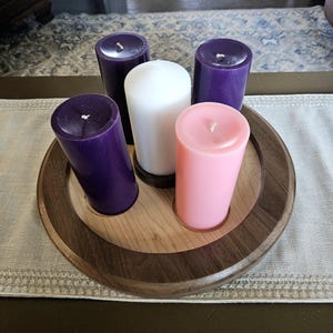 May include: A wooden candle holder with five pillar candles. The candles are purple, white, and pink. The holder is round with a light wood center and a dark wood rim. The candles are arranged in a circle.
