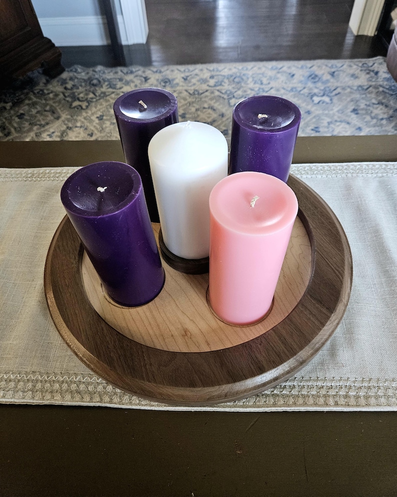 May include: A wooden candle holder with five pillar candles. The candles are purple, white, and pink. The holder is round with a light wood center and a dark wood rim. The candles are arranged in a circle.