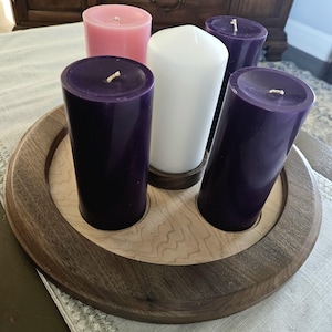 May include: A wooden candle holder with five pillar candles. The candles are purple, pink, and white. The holder is round with a light wood center and a dark wood border. The candles are arranged in a circle.