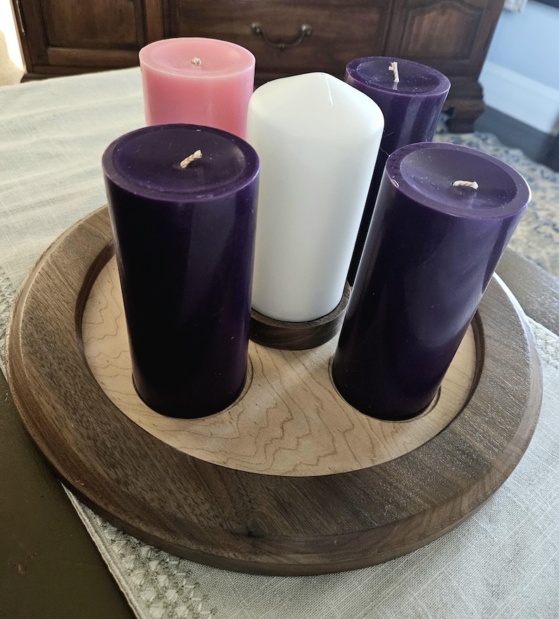 May include: A wooden candle holder with five pillar candles. The candles are purple, pink, and white. The holder is round with a light wood center and a dark wood border. The candles are arranged in a circle.
