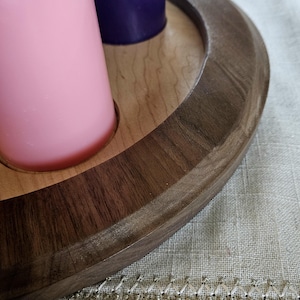 May include: A wooden candle holder with a dark brown rim and a light brown interior. It holds a pink and a purple candle. The holder is circular and sits on a textured, light-colored surface.
