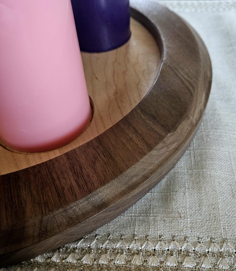 May include: A wooden candle holder with a dark brown rim and a light brown interior. It holds a pink and a purple candle. The holder is circular and sits on a textured, light-colored surface.