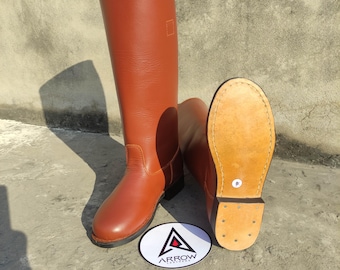 May include: A pair of brown leather riding boots. One boot is upright, the other is lying on its side, revealing the sole. The boots are tall, with a smooth finish. A white oval with the word "ARROW" is in front of the boots.