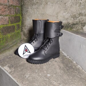 May include: A pair of black leather combat boots with laces and buckle straps. The boots have a round toe and a thick sole. A white circular tag with the word "ARROW" and a triangle logo is next to the boots.