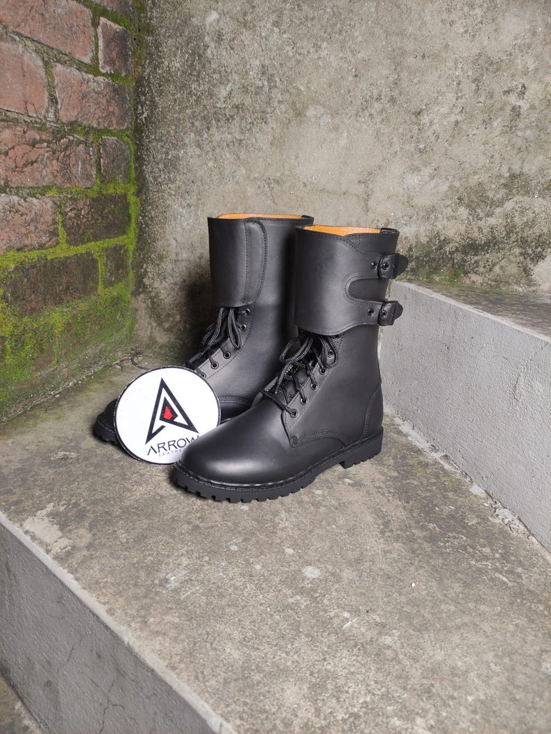 May include: A pair of black leather combat boots with laces and buckle straps. The boots have a round toe and a thick sole. A white circular tag with the word "ARROW" and a triangle logo is next to the boots.