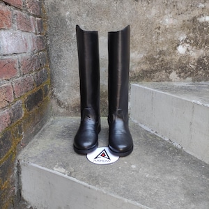 Puede incluir: Un par de botas de montar negras. Las botas están hechas de un material liso y brillante y tienen un diseño clásico. Las botas están sobre una superficie de hormigón, con un logotipo blanco y azul entre ellas.
