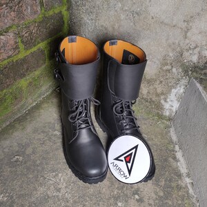 May include: A pair of black leather combat boots with laces and a buckle closure. The boots have a folded-over cuff and a round, white patch with the word "ARROW" and a logo.