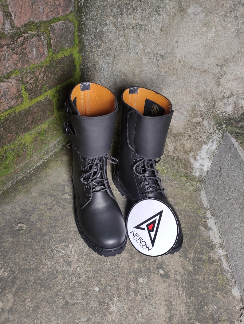 May include: A pair of black leather combat boots with laces and a buckle closure. The boots have a folded-over cuff and a round, white patch with the word "ARROW" and a logo.