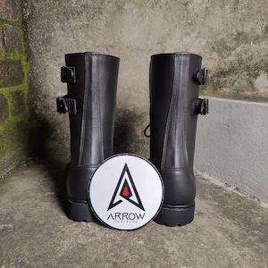 May include: A pair of black leather boots with two buckles on each boot. The boots are mid-calf height. A white, circular patch with the text "ARROW LEATHERS" and a stylized arrow logo is between the boots.