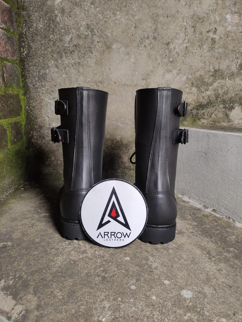 May include: A pair of black leather boots with two buckles on each boot. The boots are mid-calf height. A white, circular patch with the text "ARROW LEATHERS" and a stylized arrow logo is between the boots.