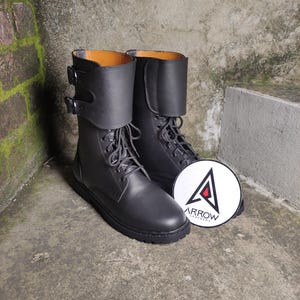 May include: A pair of black leather boots with laces and buckles. The boots have a folded-over cuff and a round logo with the text "ARROW LEATHERS". The boots are displayed on a concrete surface.