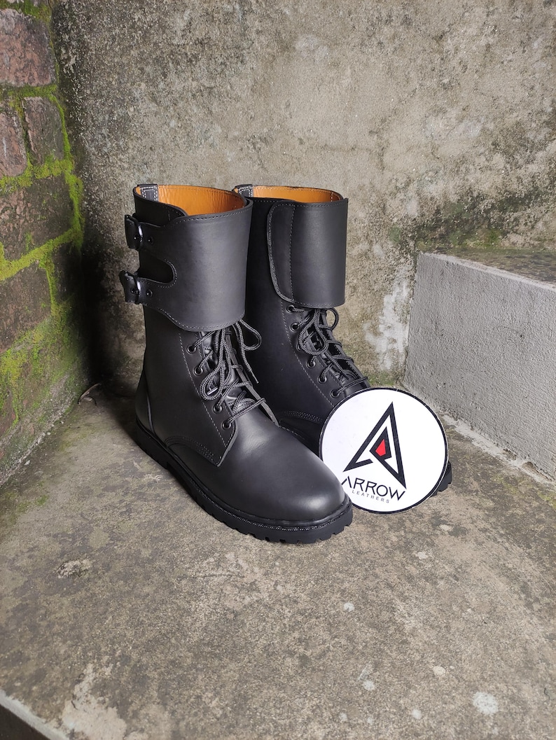 May include: A pair of black leather boots with laces and buckles. The boots have a folded-over cuff and a round logo with the text "ARROW LEATHERS". The boots are displayed on a concrete surface.