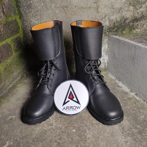 May include: A pair of black leather boots with laces and a folded-over top. The boots have a round toe and a thick sole. A white and black circular logo with the text "ARROW LEATHERS" is between the boots.