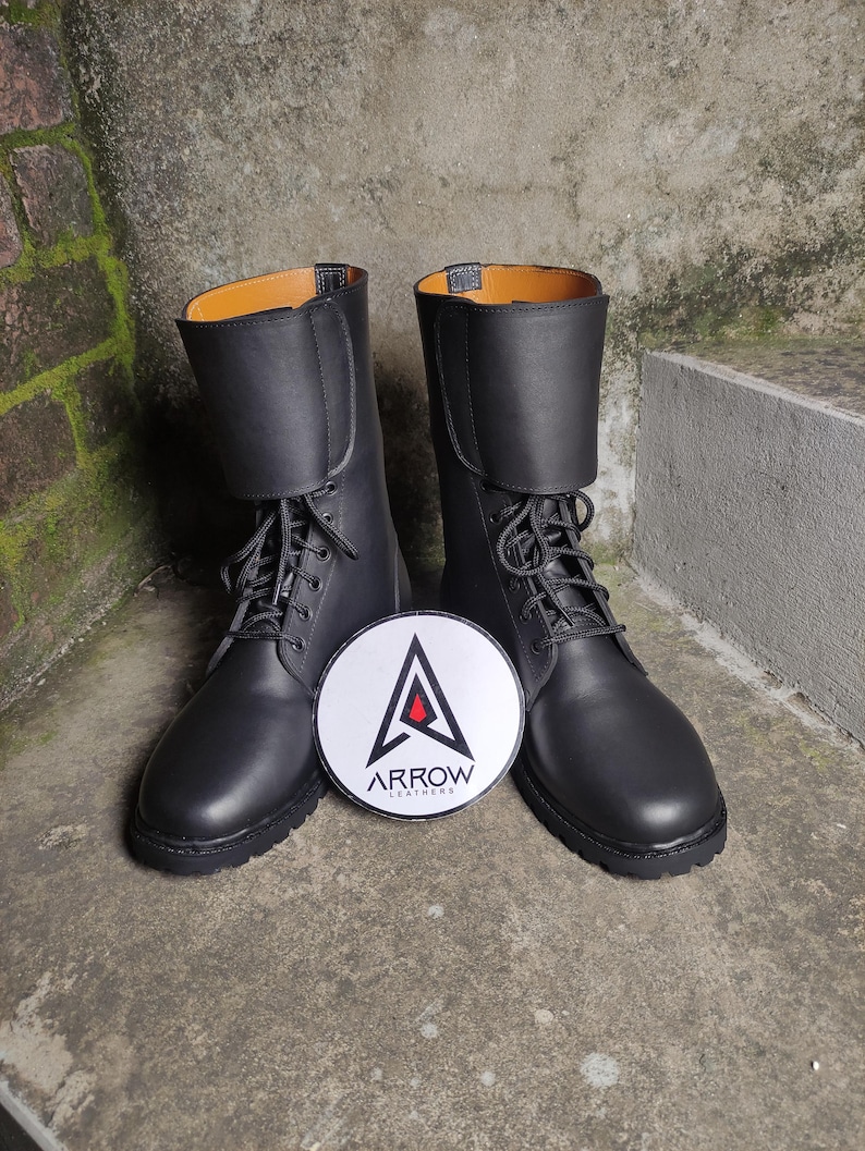 May include: A pair of black leather boots with laces and a folded-over top. The boots have a round toe and a thick sole. A white and black circular logo with the text "ARROW LEATHERS" is between the boots.