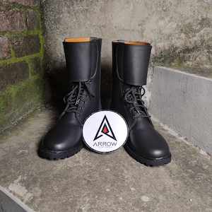 May include: A pair of black leather boots with laces and a round logo in front. The boots have a mid-calf height and a folded-over top. The logo is white with a red triangle and the text "ARROW LEATHERS".
