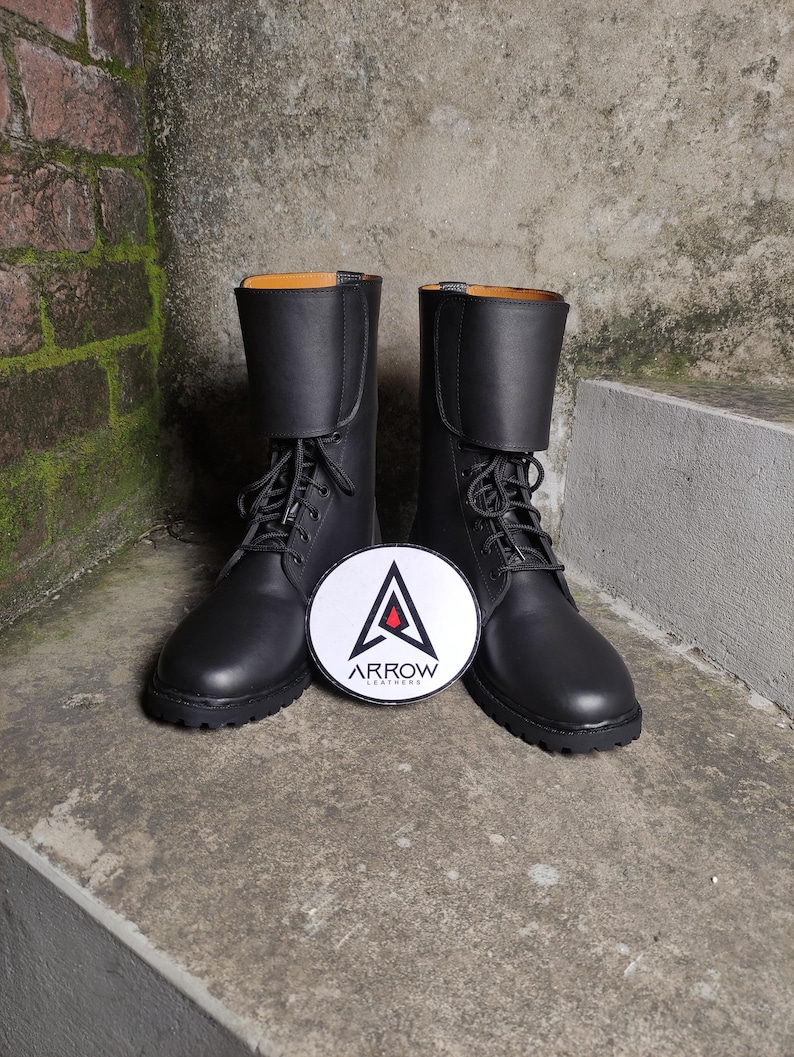 May include: A pair of black leather boots with laces and a round logo in front. The boots have a mid-calf height and a folded-over top. The logo is white with a red triangle and the text "ARROW LEATHERS".
