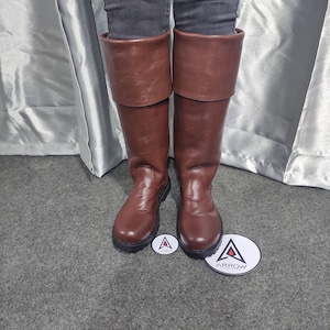 May include: Brown leather knee-high boots with folded-over tops. The boots have a smooth texture and a dark brown color. The boots are shown with a gray background and a white circle with the word "ARROW" on it.