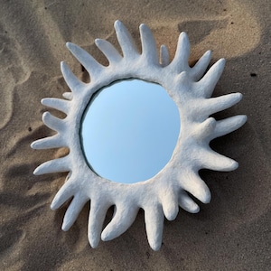 Peut inclure: Miroir blanc en forme de soleil avec une surface réfléchissante ronde. Le cadre du miroir présente un design texturé et épineux. Le miroir est posé sur une surface sablonneuse.