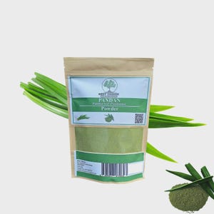 May include: A kraft paper bag of pandan powder with a green and white label. The label reads "Pandan Pandan leaf (Pandanis) Powder". Fresh green pandan leaves are arranged around the bag, and a small pile of powder is in the lower right corner.
