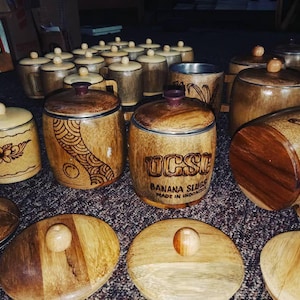 May include: A selection of handcrafted wooden containers with lids. Some containers feature etched designs, while others display the text "UCSC BANANA SLUGS MADE IN INDONESIA." The lids have small wooden knobs.