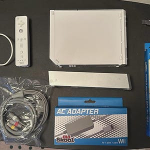 Könnte beinhalten: Eine weiße Nintendo Wii-Konsole mit passender Fernbedienung und Nunchuk-Controller, zusammen mit einer Sensorleiste, einem Netzteil und AV-Kabeln. Auf der Netzteilbox steht "Old Skool" und "AC ADAPTER". Ein USB-Stick ist ebenfalls enthalten.