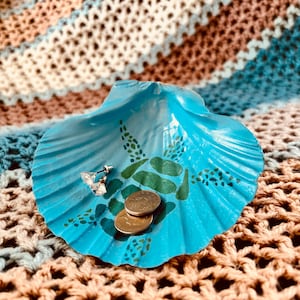 May include: A turquoise painted seashell trinket dish with green accents, holding a silver butterfly earring and two gold coins. The dish sits on a crocheted blanket with brown, beige, and blue stripes.