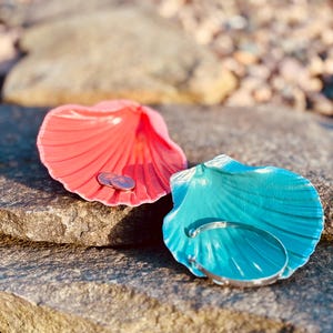 May include: Two seashell-shaped dishes, one coral and one turquoise, sit on a stone surface. The coral shell holds two coins, while the turquoise shell features a silver bracelet. The shells have a glossy finish.