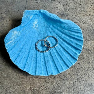 May include: A vibrant blue seashell-shaped dish holding two silver rings. The dish has a textured finish and is placed on a grey surface. The rings include a plain band and a twisted rope design.
