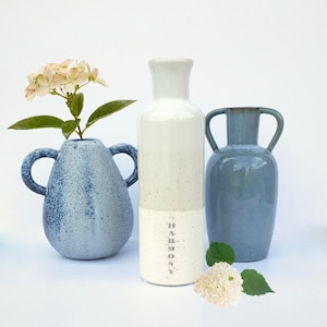 Könnte beinhalten: Drei Keramikvasen in Blau- und Weißtönen. Die Vase links ist ein gesprenkeltes Blau mit Henkeln und hält ein Blumenarrangement. Die mittlere Vase ist weiß mit dem Aufdruck "HARMONY". Die Vase rechts ist blau mit Henkeln.