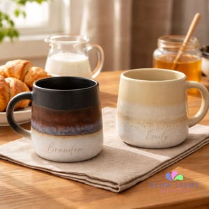 May include: Two ceramic mugs, each with a name, "Brandon" and "Emily", are displayed on a neutral napkin. The mugs feature a gradient design, one in black, brown, and cream, the other in cream and beige. A jar of honey and croissants are in the background.
