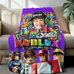 May include: A purple Roblox-themed blanket featuring a colorful graphic of a smiling character wearing a headset and holding a game controller. The blanket also displays the Roblox logo and several cartoon avatars.