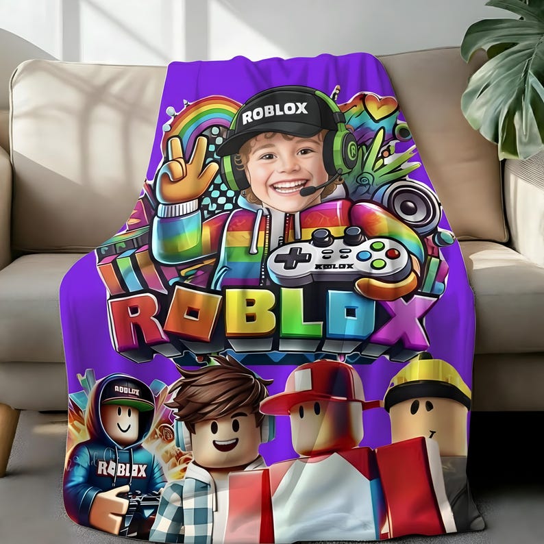 May include: A purple Roblox-themed blanket featuring a colorful graphic of a smiling character wearing a headset and holding a game controller. The blanket also displays the Roblox logo and several cartoon avatars.