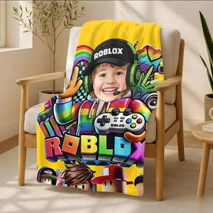 May include: A yellow blanket featuring a colorful graphic of a smiling child wearing a Roblox hat and headset, holding a game controller. The blanket has the Roblox logo in large, colorful letters.