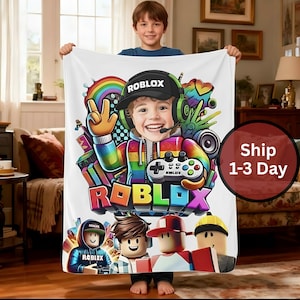 May include: White blanket featuring a colorful Roblox design. The design includes a smiling child wearing a Roblox hat and headset, a rainbow, and the Roblox logo. The blanket is held by a child.