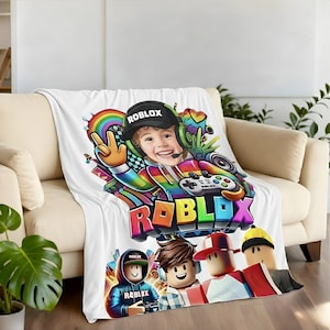 May include: White blanket featuring a colorful graphic of a child wearing a Roblox hat and holding a game controller. The blanket also displays the Roblox logo and several cartoon characters.