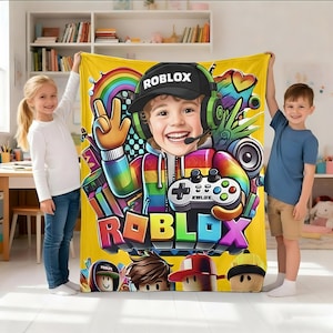 May include: A yellow fleece blanket featuring a central graphic of a smiling child wearing a black Roblox cap and green headphones, holding a game controller. The design includes a rainbow-colored hoodie, a rainbow, and the word Roblox in bold letters.