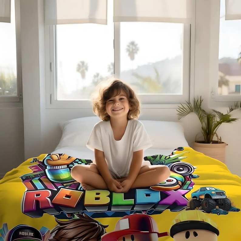 May include: A young person sits on a bed with a yellow blanket featuring the Roblox logo and characters. The blanket has a colorful design with various game elements and characters. The person is wearing a white t-shirt.
