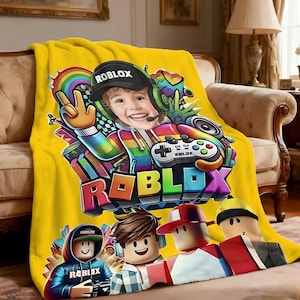 May include: A yellow fleece blanket featuring a colorful Roblox design. The design includes a smiling child wearing a Roblox cap and headset, a rainbow, and the Roblox logo. The blanket also features several Roblox characters.