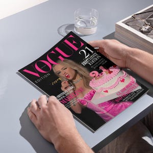 May include: A person holding a copy of Vogue magazine. The cover features the text "VOGUE EDITION" in pink, with "21 BIRTHDAY EDITION" and a photo of a woman in pink. A pink cake is also featured on the cover.