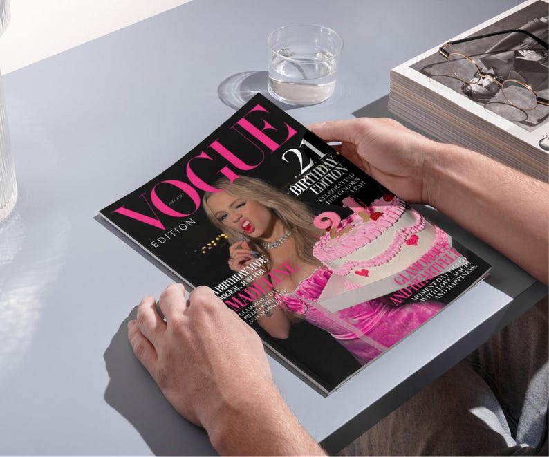 May include: A person holding a copy of Vogue magazine. The cover features the text "VOGUE EDITION" in pink, with "21 BIRTHDAY EDITION" and a photo of a woman in pink. A pink cake is also featured on the cover.