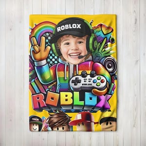 May include: Yellow blanket featuring a colorful graphic of a smiling child wearing a Roblox hat and headset, holding a game controller. The word "ROBLOX" is prominently displayed in 3D letters. A rainbow and other game-related elements complete the design.