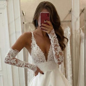 Può includere: Un abito da sposa bianco con scollo a V profondo e gonna ampia. Il corpetto presenta dettagli in pizzo intricati e le maniche lunghe sono in pizzo delicato. Altri abiti da sposa sono visibili sullo sfondo.