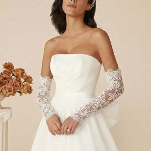 May include: A white strapless wedding dress with a fitted bodice and a flowing skirt. The dress is complemented by long, delicate lace gloves. A bouquet of orange roses is in a white vase on a pedestal.