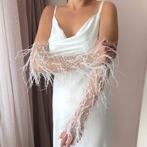 May include: A white satin slip dress with spaghetti straps. The dress is paired with sheer, long gloves adorned with white feathers and small, sparkling embellishments. The gloves add a touch of elegance to the simple dress.