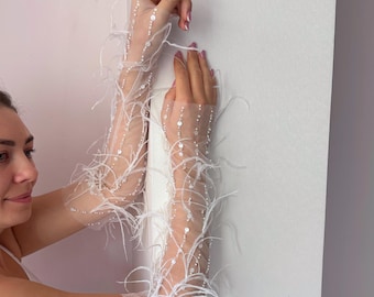 May include: Sheer white gloves adorned with delicate white feathers and small, sparkling embellishments. The gloves extend from the fingertips to the upper arm, creating an elegant and ethereal look. The gloves are a bridal or formal wear accessory.